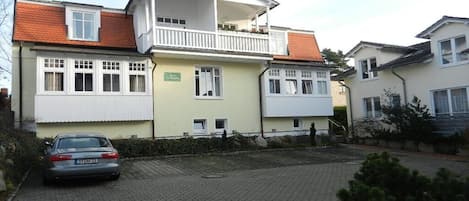Um edifício residencial de dois andares com varanda, um carro estacionado na frente e um acesso pavimentado.