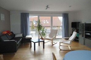 A modern living room with a sofa, a coffee table, a rocking chair, a television, and a balcony with a view of houses. - Holiday apartment with 1 bedroom (Ostbevern)