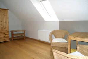 A room with a sloped ceiling, a wooden cabinet, a wicker chair, a small table, and a radiator. - Holiday apartment with 1 bedroom (Ostbevern)