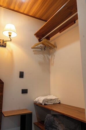 Canto de um quarto com teto de madeira, luminária na parede, um cabideiro e uma toalha dobrada em uma prateleira.