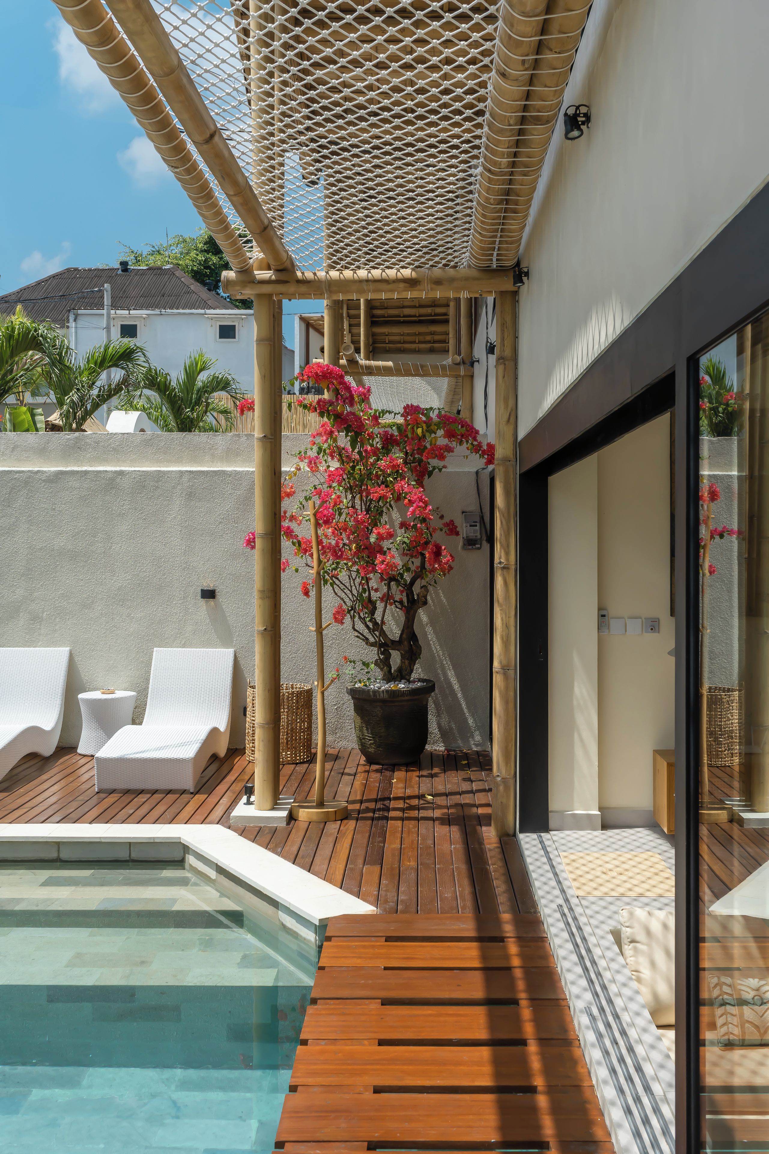 Zona junto a la piscina con una terraza de madera, sillas de descanso blancas, una planta en maceta con flores rosadas y una estructura de pérgola.
