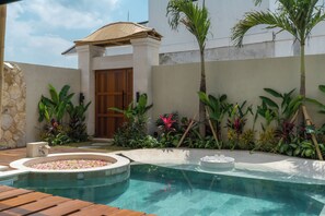 A swimming pool with a wooden deck, surrounded by tropical plants and a stone wall. - Villa Tigana (Pererenan)