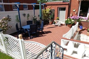 Eine Terrasse mit rundem Tisch und Stühlen, umgeben von Kübelpflanzen und einem weißen Zaun.