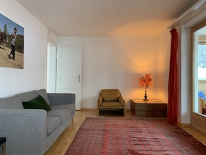 Un salon avec un canapé gris, un tapis à motifs, une petite table avec une lampe et une porte donnant sur l’extérieur.