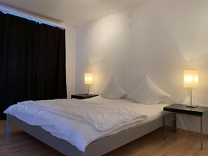 Une chambre d’hôtel moderne avec un grand lit, deux tables de chevet avec des lampes et une vue sur le ciel par une fenêtre munie de rideaux.