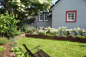 Ein Garten mit einer Bank, ein Haus mit rotem Fensterrahmen und blühenden weißen Blumen.