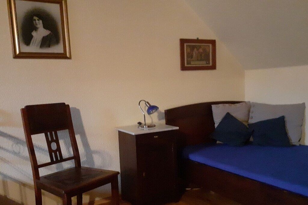 Een slaapkamer met een bed, een stoel, een bureau met een lamp en twee foto's in lijsten aan de muur.