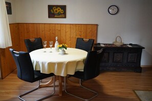 Ein Esstisch mit weißer Tischdecke, vier schwarze Stühle, eine Vase mit gelben Blumen und eine Uhr an der Wand.
