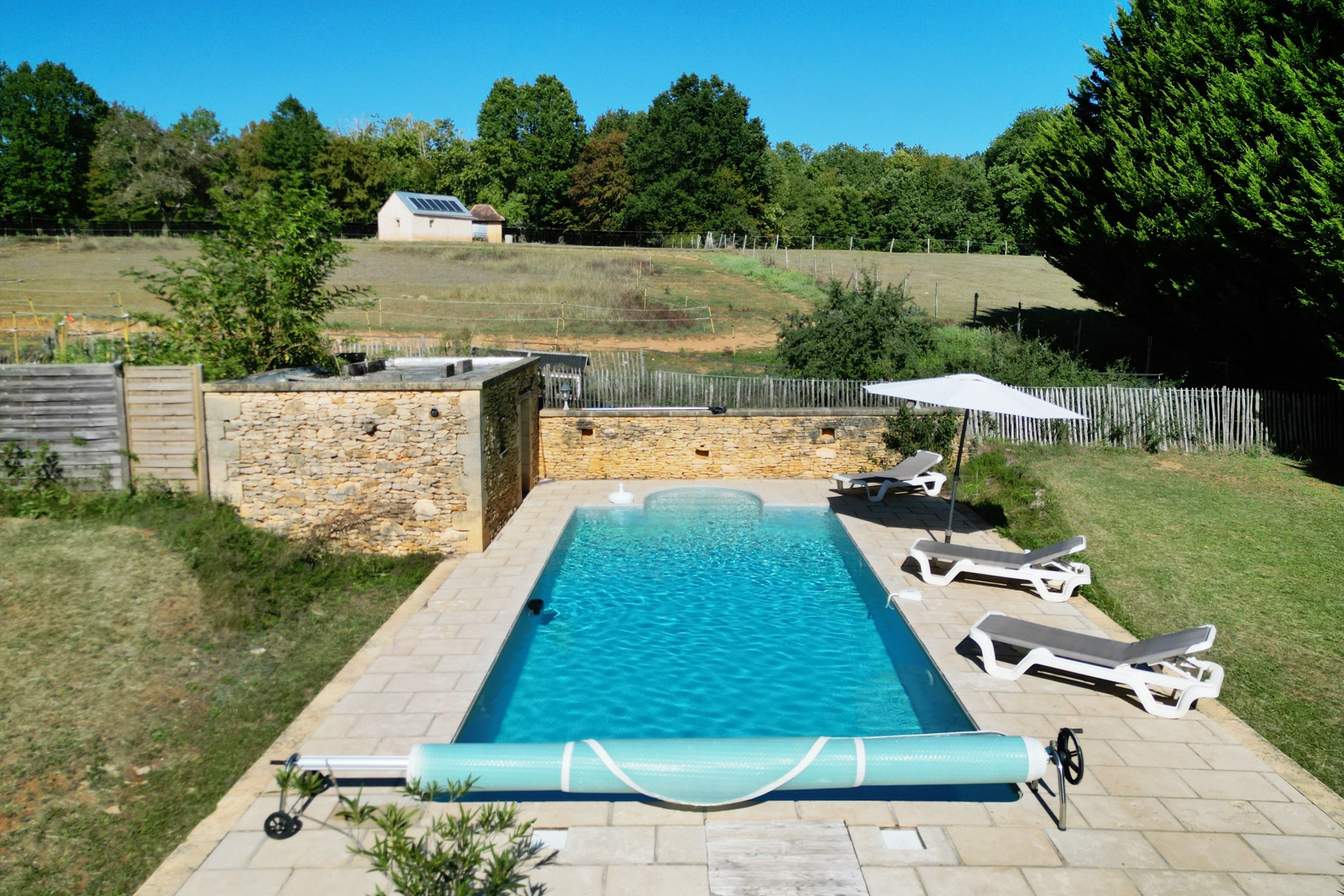 Una piscina rectangular con un muro de piedra en un lado y un césped con árboles y una valla en el fondo.