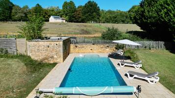 Una piscina rectangular con un muro de piedra en un lado y un césped con árboles y una valla en el fondo.