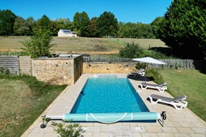 Una piscina rettangolare con un muro in pietra su un lato e sullo sfondo un'area verde alberata e recintata.