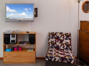 A room with a TV mounted on the wall, a wooden bookshelf, a patterned armchair, and a floor lamp. - Colette 42 Appartement/Fewo, Dusche oder Bad, WC 2-Zimmerwohnung by Interhome (Davos)