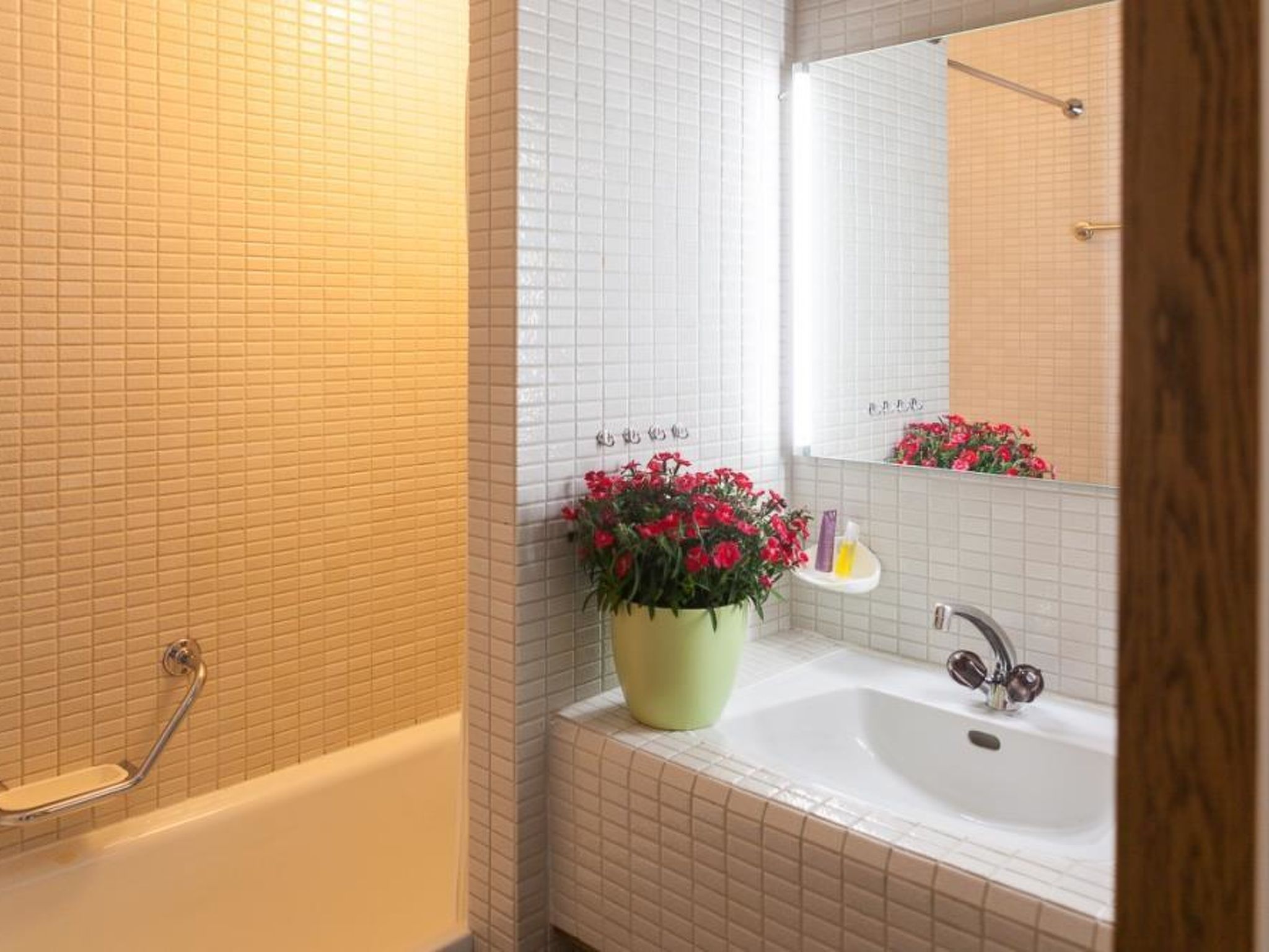 Et badeværelse med et hvidt badekar, en vask med et spejl og en potteplante med røde blomster.
