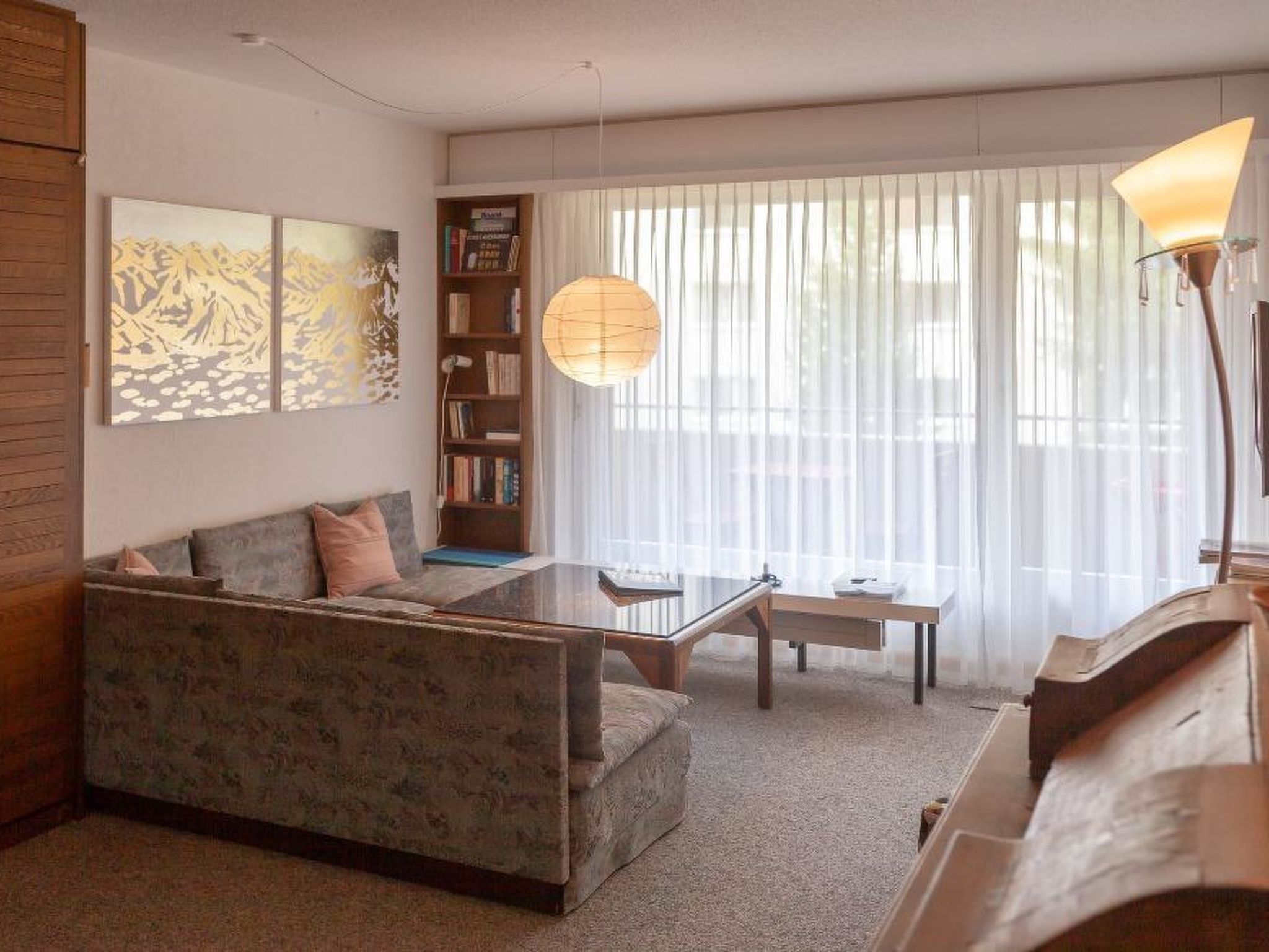 A living room with a sofa, a coffee table, a bookshelf, and a large window with sheer curtains.