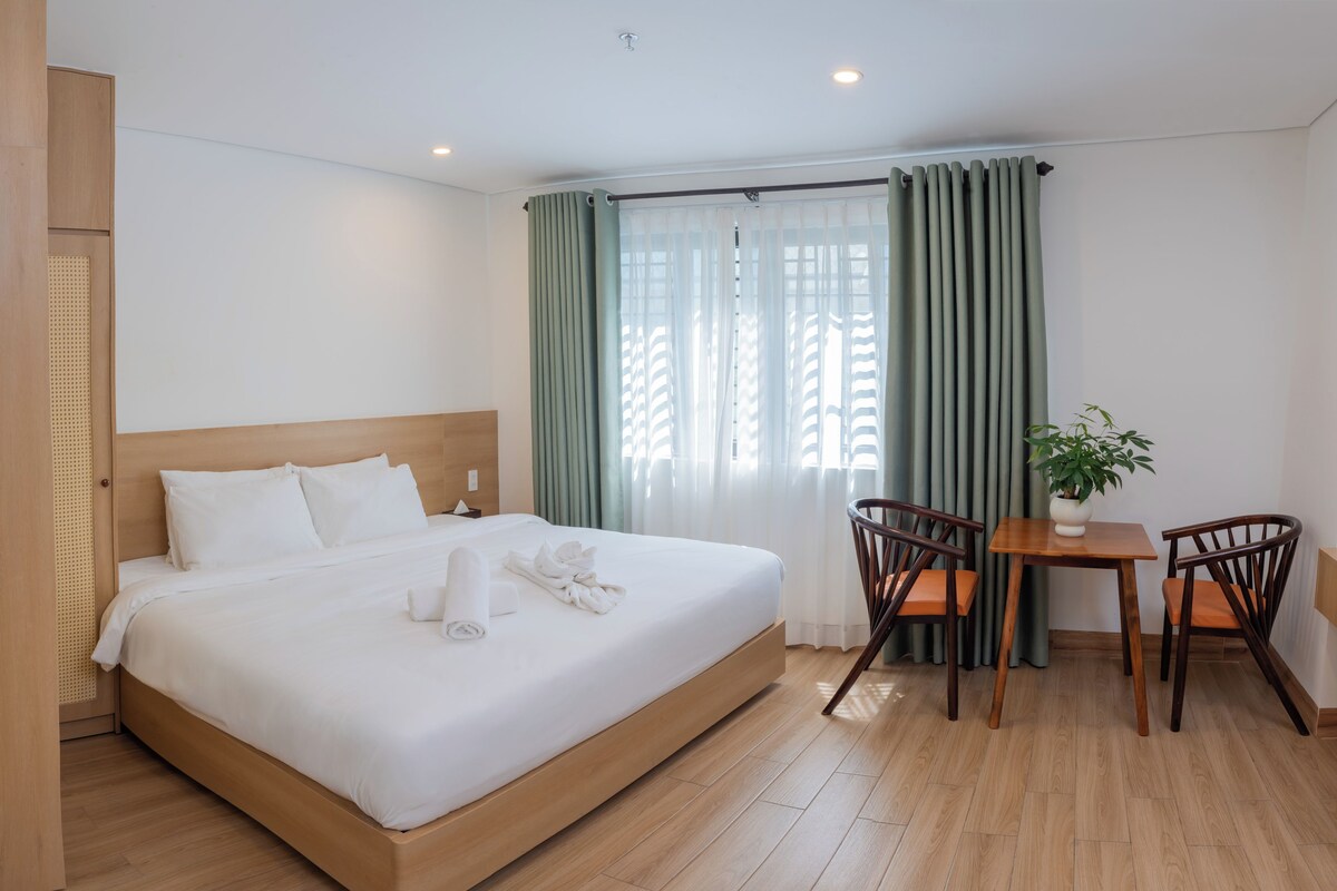 Habitación de hotel con una cama grande, cabecera de madera, una mesita con sillas y una planta en maceta.