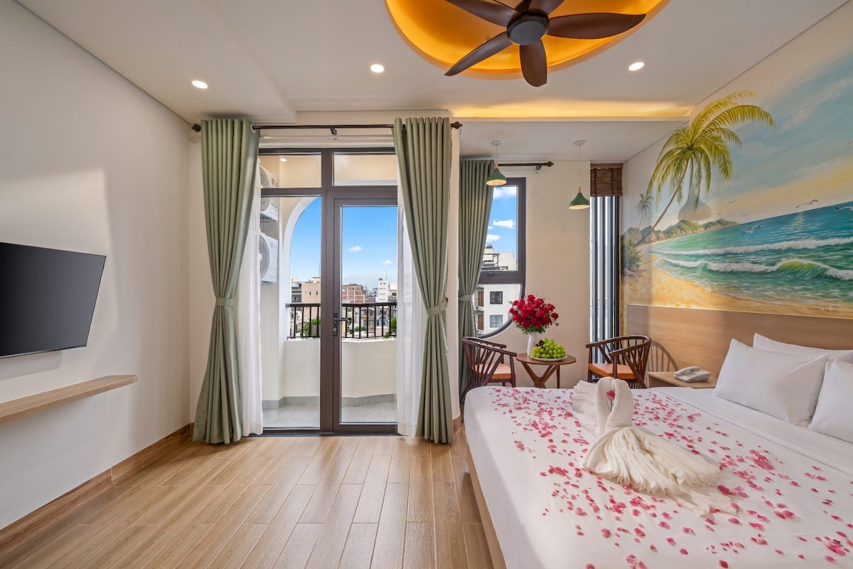 Quarto de hotel com cama, televisão, varanda com cortinas, pintura de uma praia e ventilador de teto.