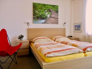 A bedroom with a wooden bed, a red chair, a small bedside table with a lamp, and a painting of a landscape. - Uf dr Egga 22 Appartement/Fewo, Dusche oder Bad, WC 2-Zimmerwohnung by Interhome (Davos)