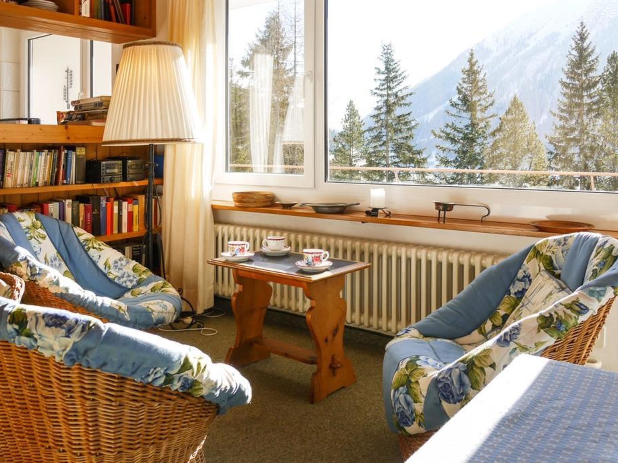 A cozy room with a large window, a bookshelf, two wicker chairs, a small table with cups, and a radiator.
