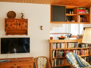 Un salon avec un téléviseur, des meubles en bois, une bibliothèque et, au fond, un coin cuisine.