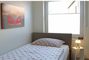 Een enkel bed met een bedsprei met dessin, een aan de muur hangend kunstwerk, een bedlampje en een raam met jaloezieën.