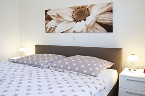 En pænt redt seng med stjernebetrukket sengetøj, et blomsterbillede på væggen og senelamper.
