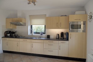 A kitchen with light wooden cabinets, a stainless steel microwave, and a window with blinds. - Holiday home with 1 bedroom (Julianadorp aan Zee)