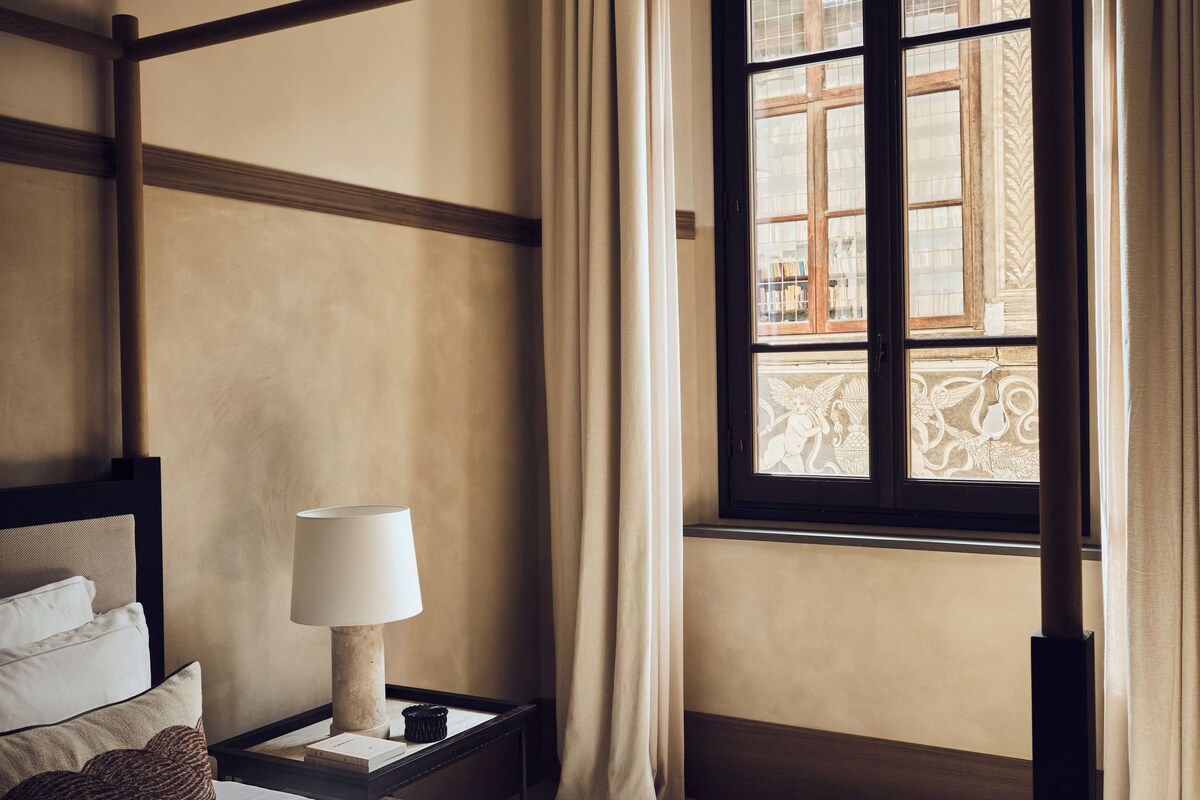 A room with a four-poster bed, a bedside table with a lamp, and a window with curtains.