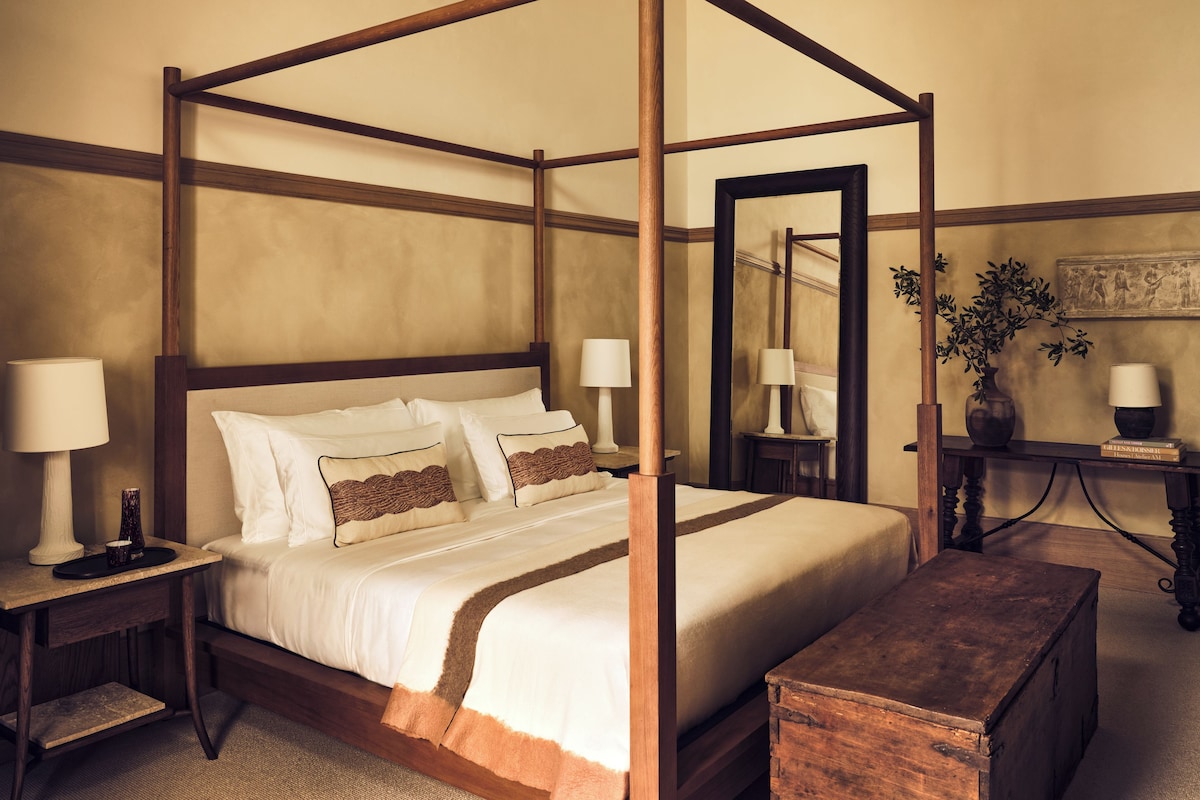 A four-poster bed with white linens and brown accents, flanked by nightstands with lamps, a wooden bench, and a mirror reflecting the room.