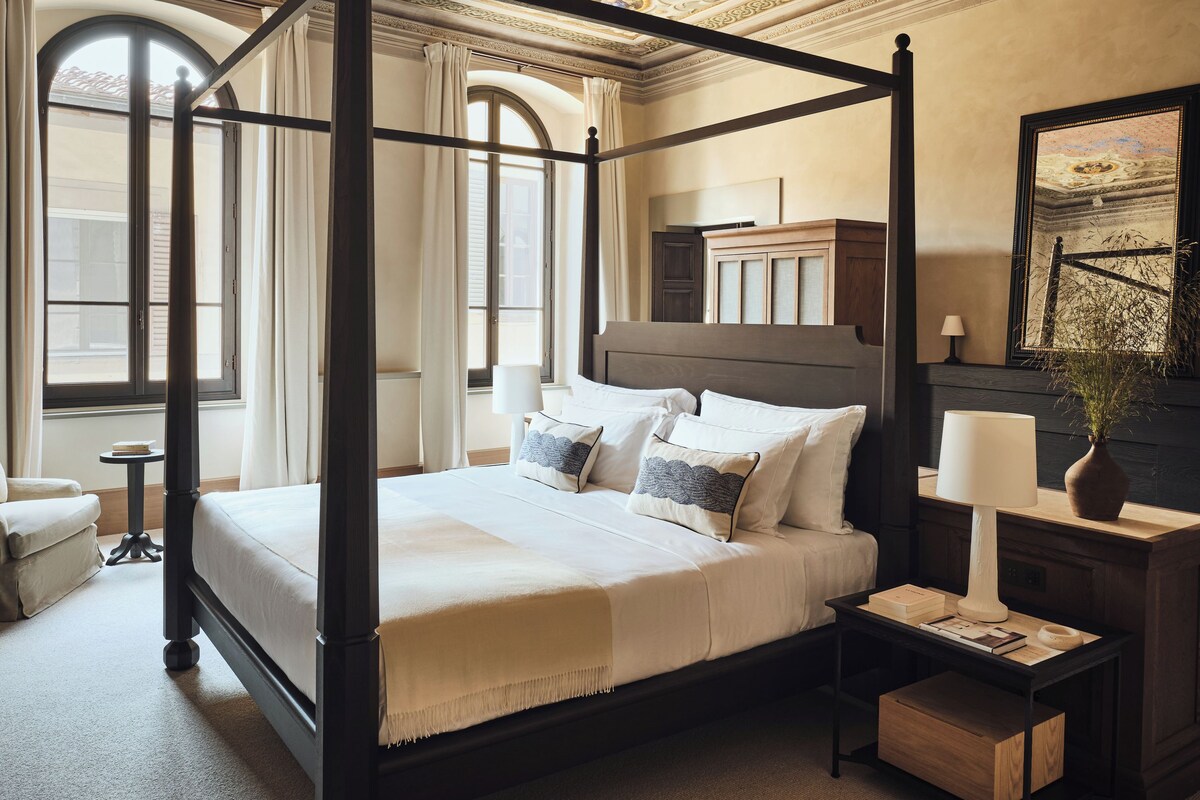 A four-poster bed with a canopy, a bedside table with a lamp, and a large window with curtains.