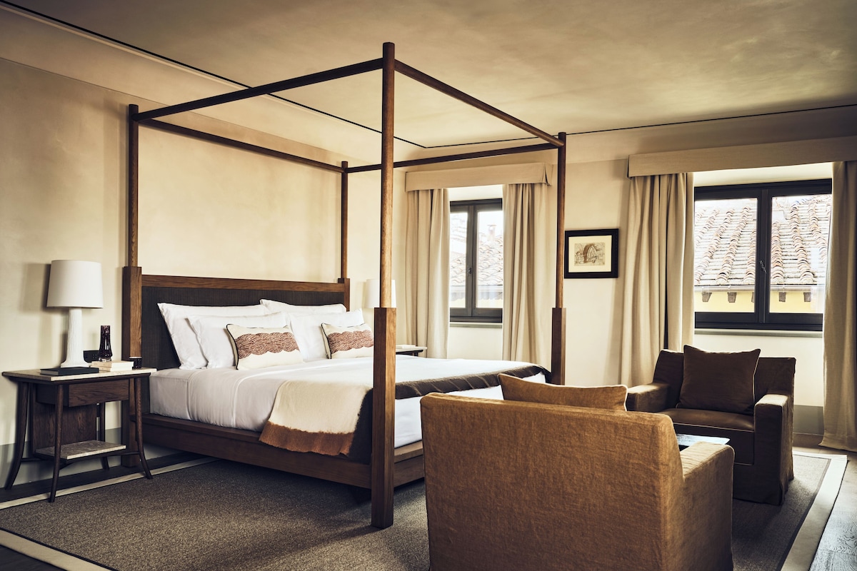 A four-poster bed with a wooden frame, a bedside table with a lamp, a chair, and a window with curtains.