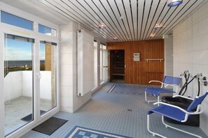 Een gang met een glazen deur naar een balkon, een houten wand en een zithoek met blauwe stoelen en een zwart bad.