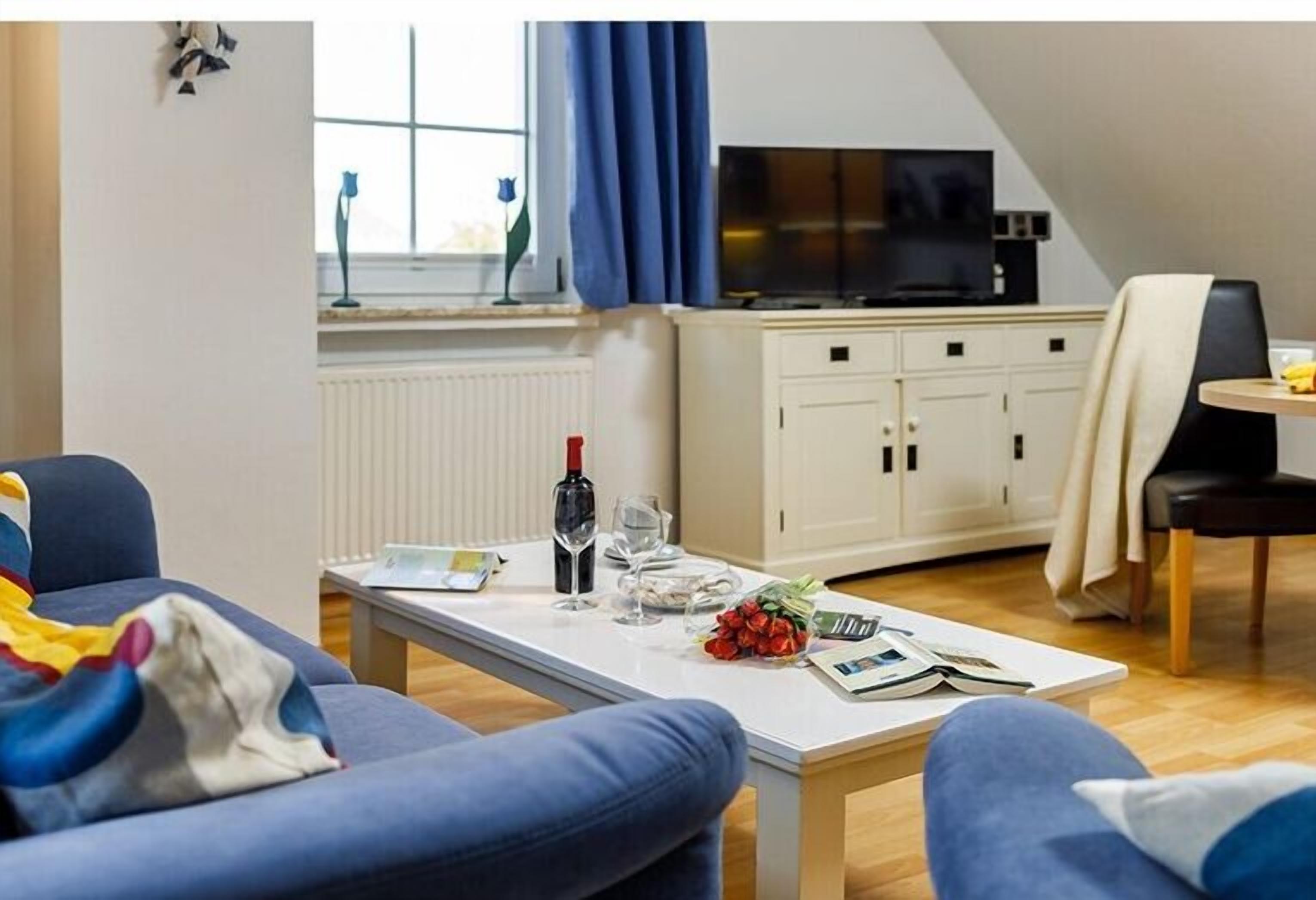 Un salon moderne avec un canapé, une table à manger agrémentée d’une bouteille de vin, de verres et d’un livre. On y trouve également un coin cuisine avec des placards et un téléviseur.