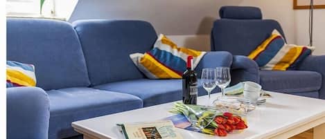 Una sala de estar con un juego de sofá, una mesa de centro con un libro, una botella, vasos y un ramo de flores.