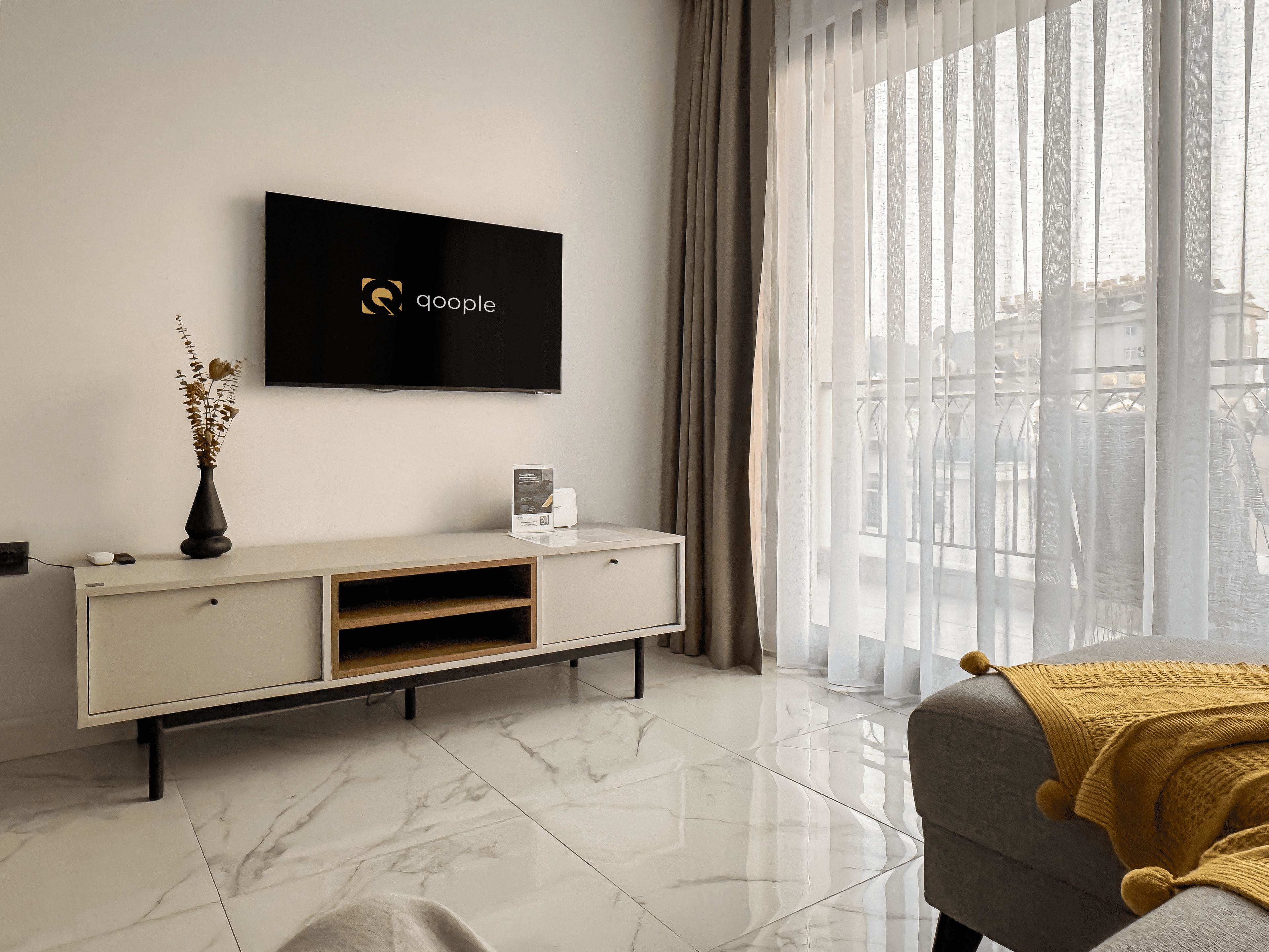 A modern living room with a flat-screen TV mounted on the wall, a white console table, and a large window with sheer curtains.