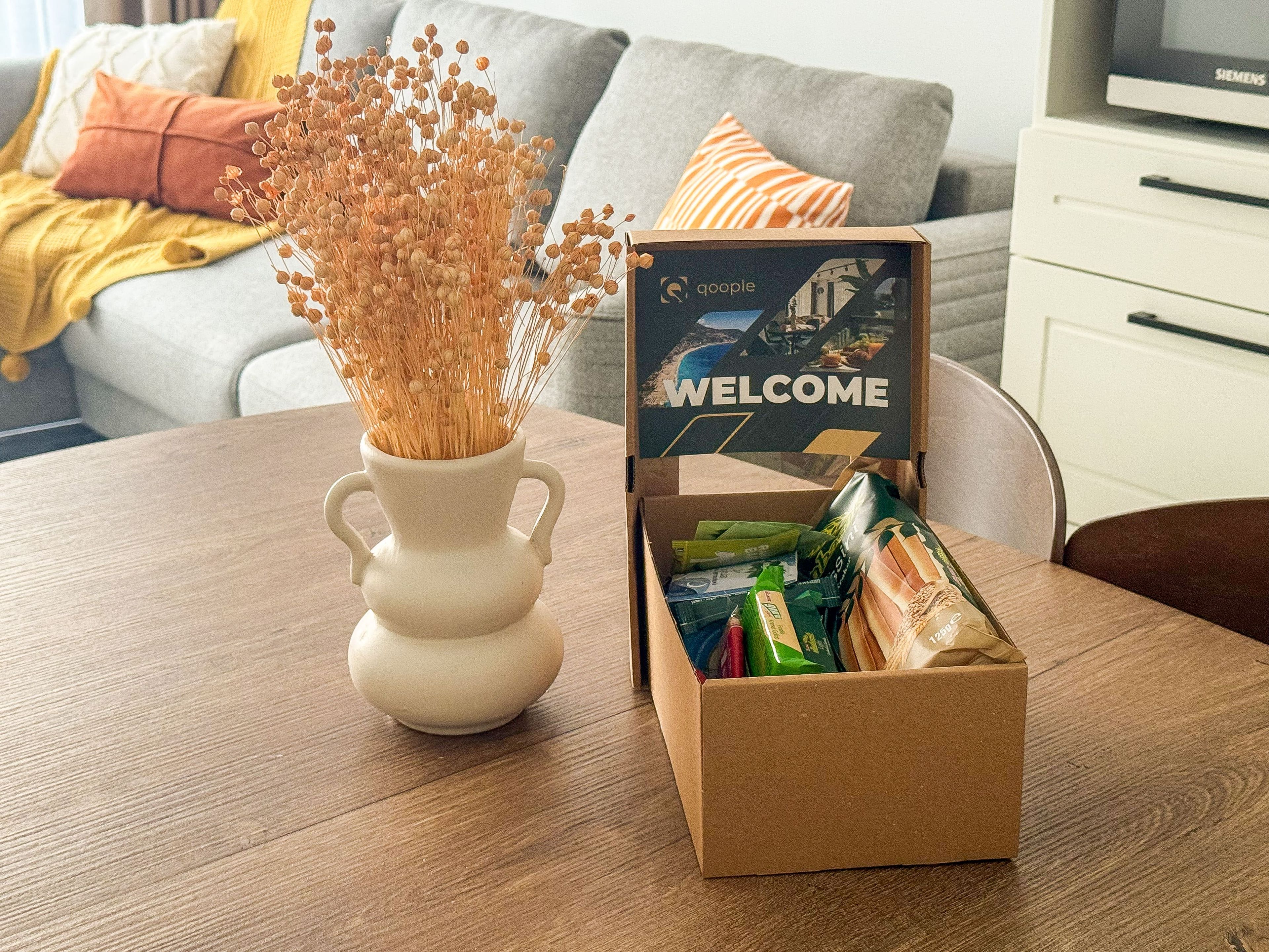 Ein Holztisch mit einer weißen Vase voller getrockneter Blumen, einer Pappe mit einem Willkommensschild und einer Tüte Snacks.