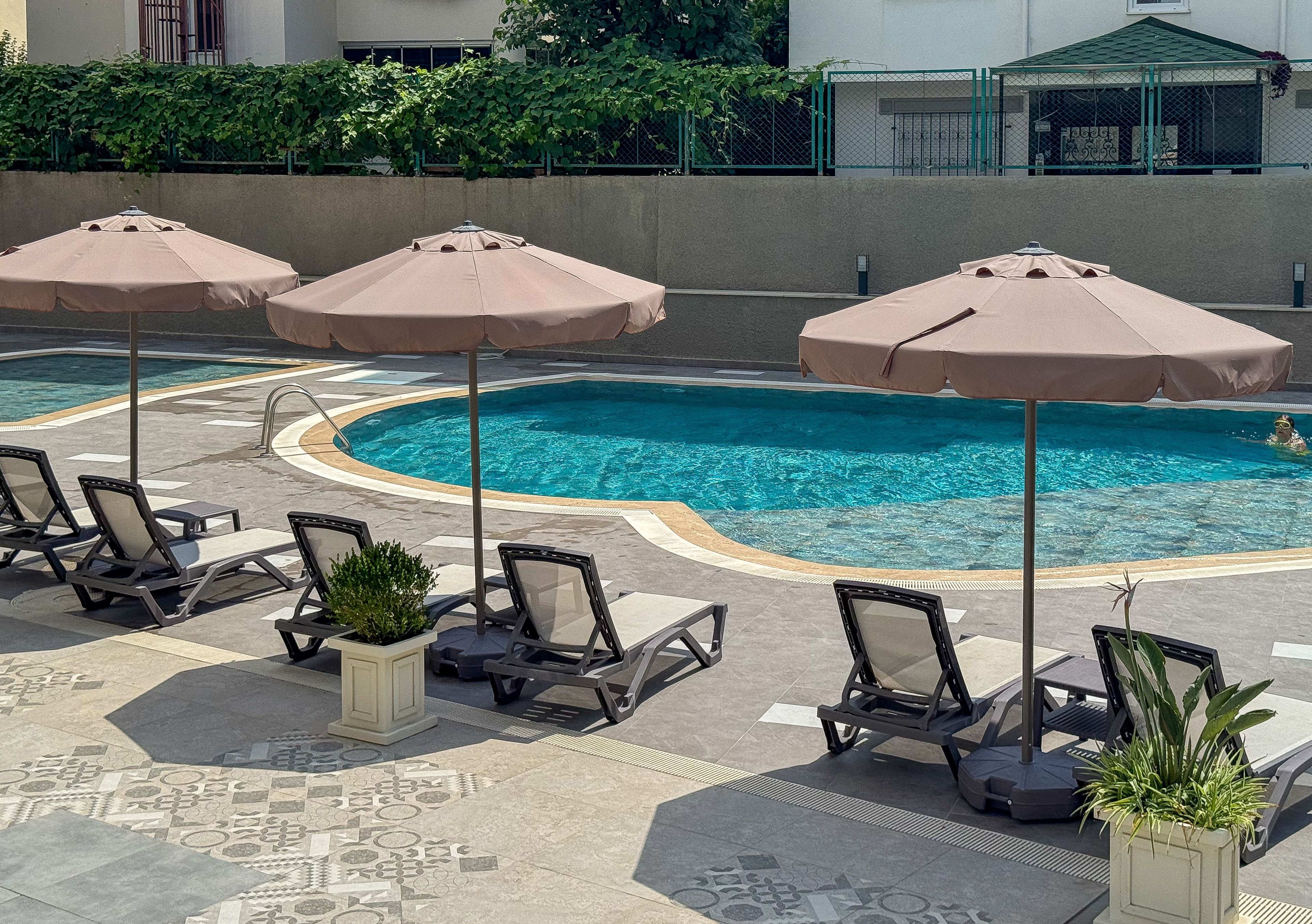 Ein Poolbereich mit Liegestühlen und Sonnenschirmen, umgeben von einer niedrigen Mauer und viel Grün.