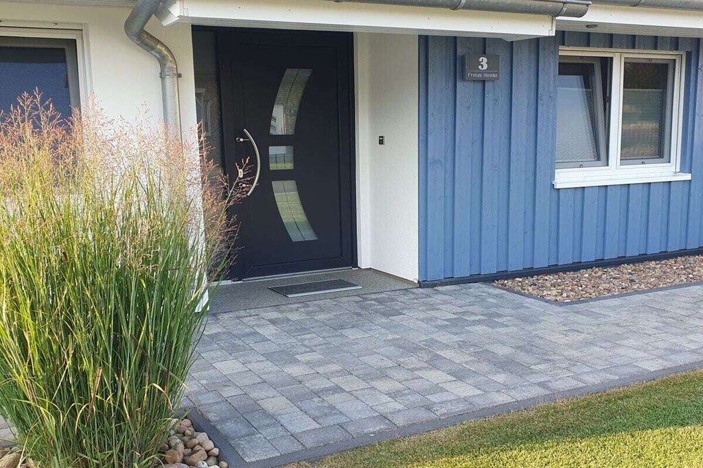 Un bâtiment moderne avec une porte noire, une façade bleue et une zone d’entrée pavée.