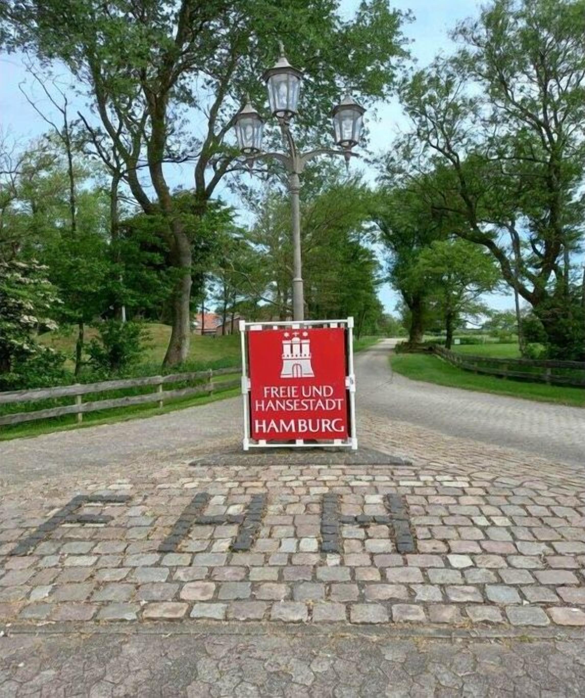 Een bord met de tekst 'FREIE UND HANSSTADT HAMBURG', een geplaveid pad en bomen op de achtergrond.