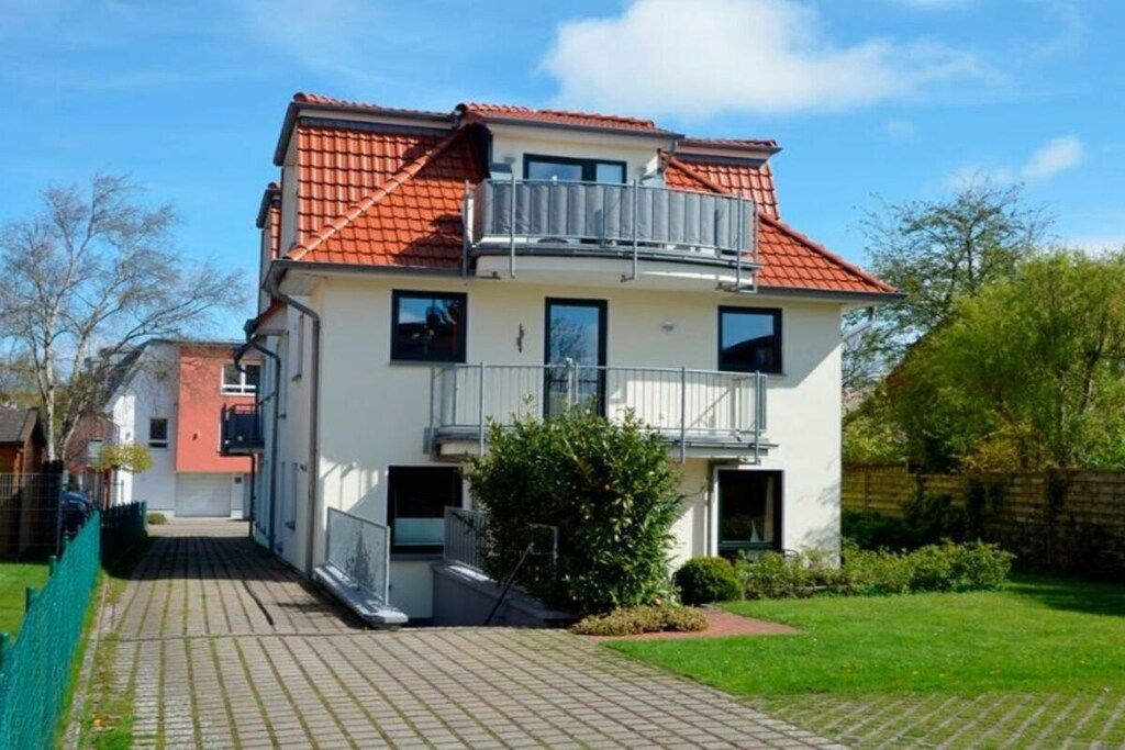 Un immeuble résidentiel à deux étages, doté d’un toit recouvert de tuiles rouges, d’un balcon à l’étage supérieur et d’une allée pavée menant à l’entrée.