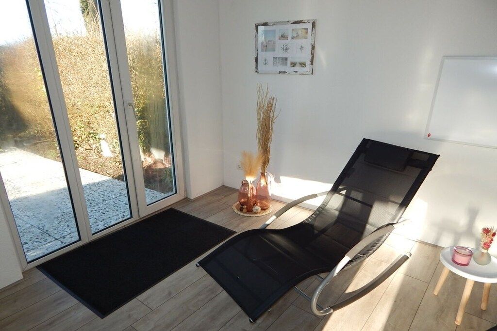 Une pièce avec une porte vitrée donnant sur l’extérieur, un tapis noir sur le sol, une chaise noire, une petite table avec un vase et un tableau blanc accroché au mur.