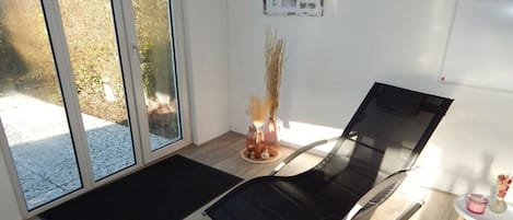 A room with a glass door leading outside, a black mat on the floor, a black chair, a small table with a vase, and a whiteboard on the wall.