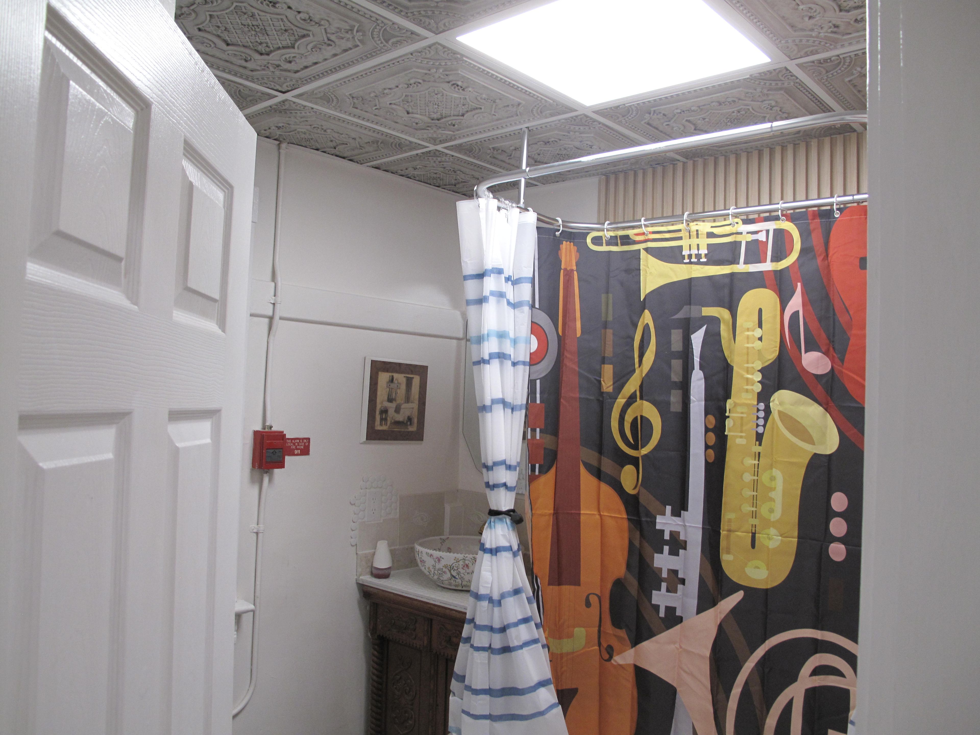 A bathroom with a shower curtain featuring musical instruments, a white door, and a wall-mounted light fixture.