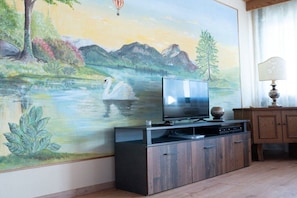 A room with a large wall mural depicting a serene landscape, a flat-screen TV on a wooden cabinet, and a lamp on a side table. - Holiday apartment with 1 bedroom (Schwangau)