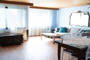 Ein Wohnzimmer mit einer Couch, einem Couchtisch und einem Esstisch mit Stühlen. Ein großes Fenster mit leichtem Vorhang und ein dekorativer Spiegel an der Wand.