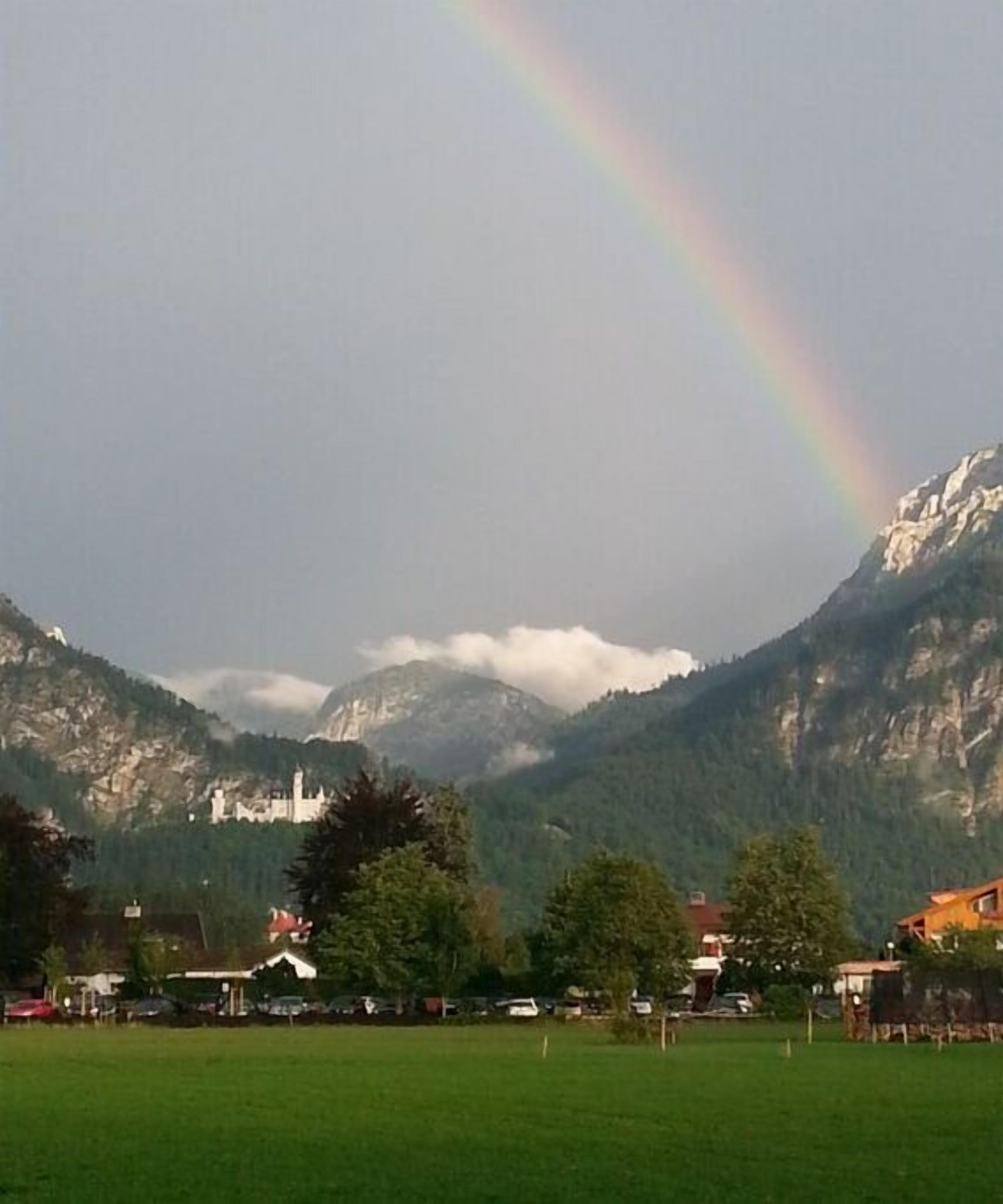 A mountain landscape with a rainbow, a castle-like building, and a green field with trees and buildings.