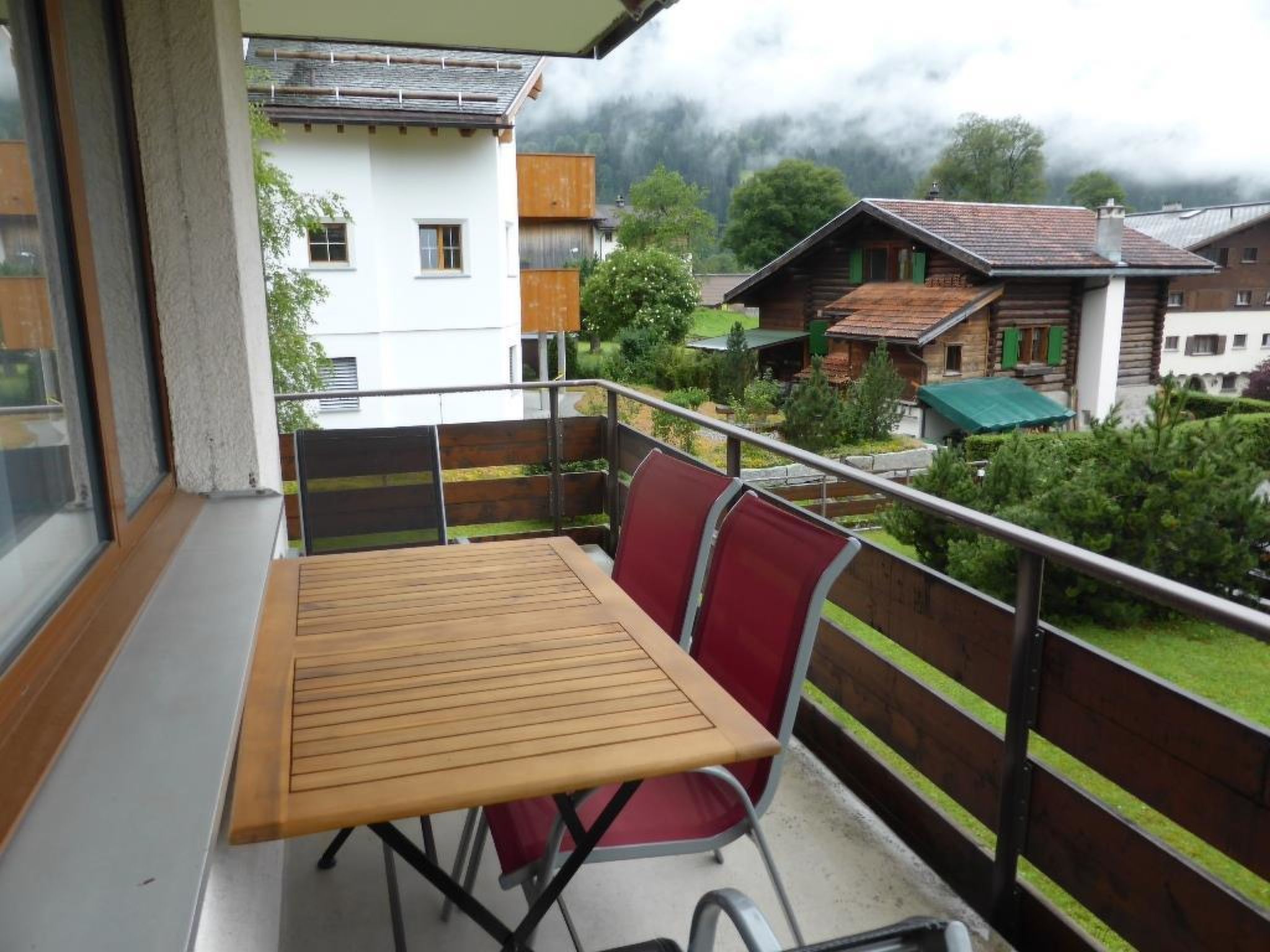 Een balkon met een houten tafel en rode stoelen, met uitzicht op een woongebied met huizen en groen.