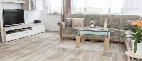 Een moderne woonkamer met een bank, een glazen salontafel, een flatscreen televisie en een witte kast.