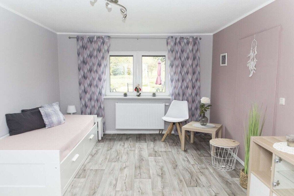 A bedroom with a bed, a chair, a small table, a radiator, and a window with curtains.