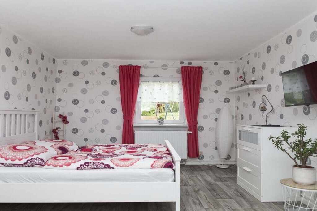 Quarto com cama, penteadeira, radiador, janela com cortinas e uma planta.