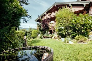 A wooden building with a balcony, surrounded by greenery and a pond. - Holiday apartment with 2 bedrooms (Ruhpolding)